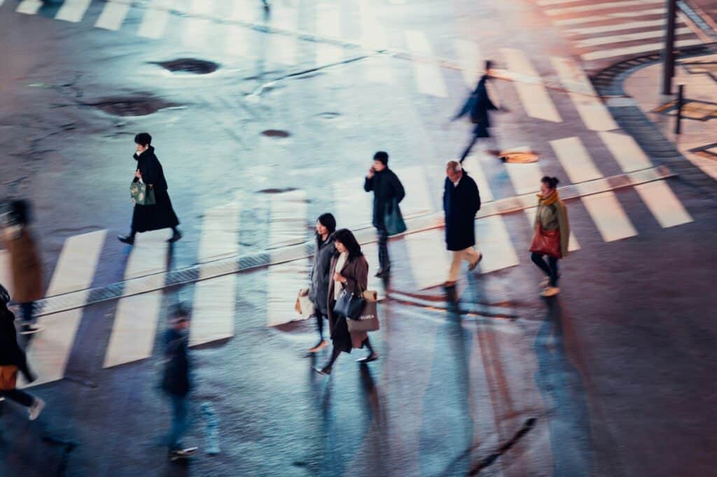 A group of people walk across a city street at a crosswalk, captured from above. The street is wet, and the scene is slightly blurred.