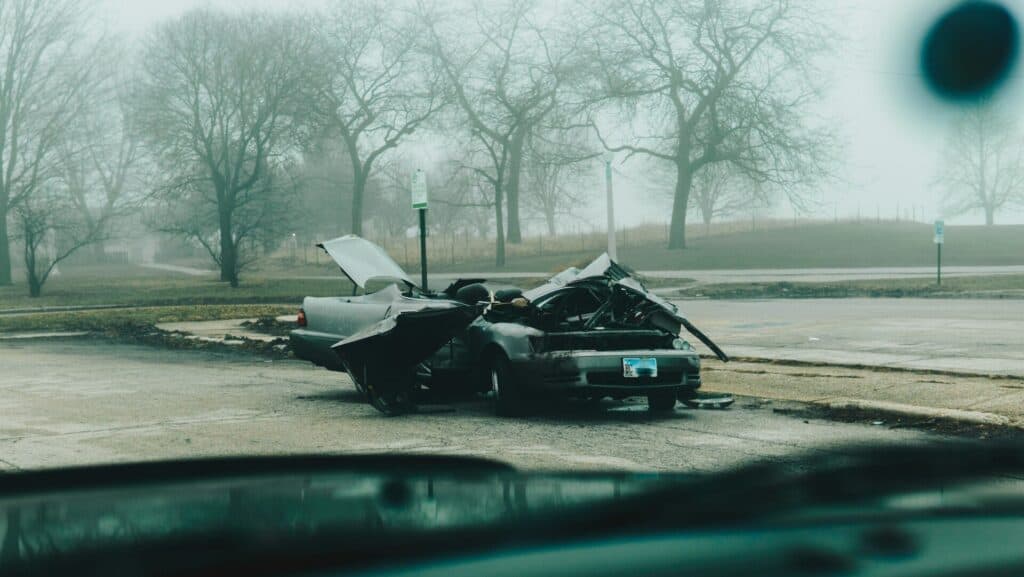 A heavily damaged car is parked by the roadside with its hood open. Trees and a foggy background are visible.