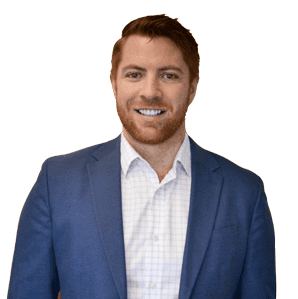A man with short red hair and a beard, wearing a blue blazer and white shirt, smiles at the camera against a plain background.